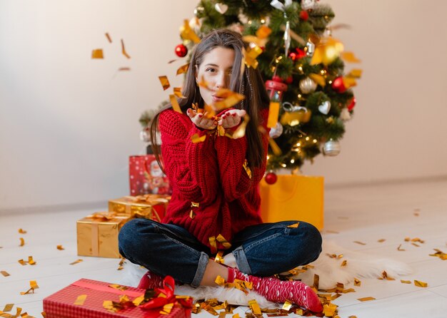 Jolie femme en pull rouge assis à la maison à l'arbre de Noël jetant des confettis dorés entouré de cadeaux et coffrets cadeaux