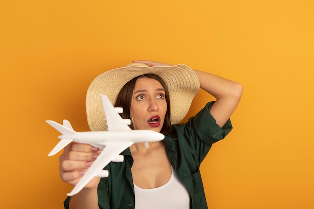 Jolie femme de race blanche anxieuse avec chapeau de plage détient avion modèle à côté sur orange