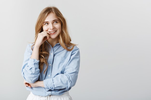 Jolie femme de race blanche aux cheveux blonds, regardant dans le coin supérieur droit, souriant curieusement et touchant la lèvre