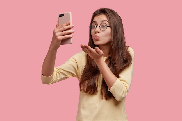 Jolie femme de race blanche fait un baiser d'air à la caméra du téléphone portable, a un appel vidéo avec son petit ami à distance, flirte et dit au revoir