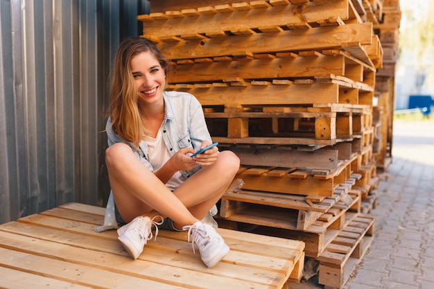 Jolie femme de race blanche avec des lèvres roses tenant un smartphone dans les mains tout en étant assis les jambes croisées