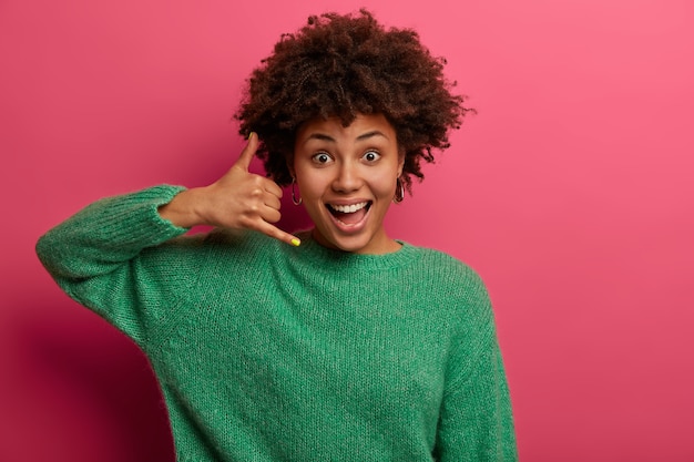 Jolie femme ravie fait signe de téléphone, dit de me rappeler, sourit joyeusement, communique avec des gestes, vêtue d'un pull vert, pose sur un mur rose. N'oublie pas d'appeler, d'être en contact