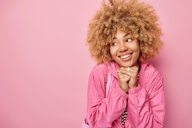 Jolie femme réfléchie a une expression heureuse garde les mains sous le menton sourit à pleines dents vêtue d'une veste concentrée loin isolé sur fond rose avec un espace vide pour votre contenu promotionnel