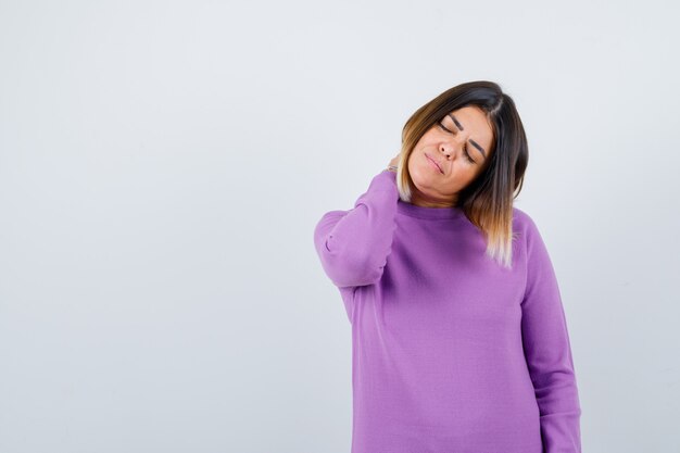 Jolie femme ressentant des douleurs au cou en pull violet et ayant l'air fatiguée, vue de face.