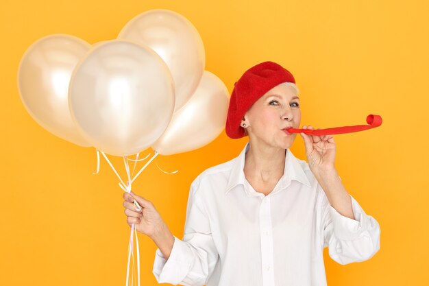 Jolie femme retraité en bonnet rouge appréciant la fête, divertissant ses petits-enfants soufflant sifflet, tenant des ballons d'hélium blanc