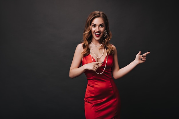 Jolie femme en robe rouge pointant vers l'endroit pour le texte Portrait intérieur de l'actrice en tenue de fête en soie posant sur fond noir