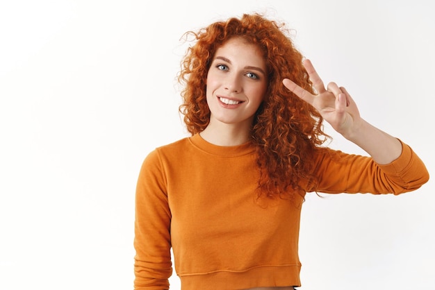 Jolie femme rousse féminine boucles naturelles montrant le signe de la victoire de la paix envoyer des vibrations positives souriant largement souhaiter à tout le monde joyeux thanksgiving debout pull orange sur fond blanc