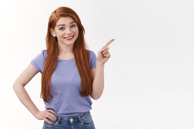 Jolie femme rousse joyeuse aux longs cheveux roux, pointant vers le coin supérieur droit, inclinant la tête et souriant, inviter le profil de visite, boutique en ligne, indiquer la bannière d'entreprise, mur blanc debout