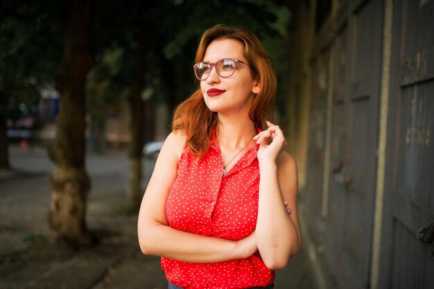 Jolie femme rousse à lunettes porter sur un chemisier rouge et une jupe en jean posant