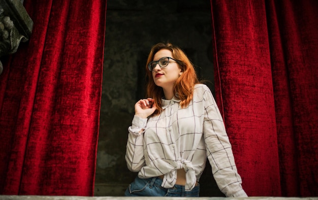 Photo gratuite jolie femme rousse portant des lunettes sur un chemisier blanc posant à l'arche de rideaux rouges ouverts