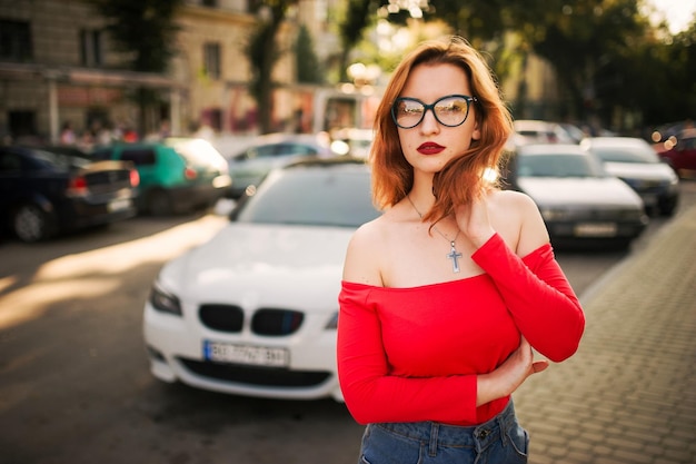 Jolie femme rousse portant des lunettes sur un chemisier rouge et une jupe en jean posant dans la rue contre une voiture de sport blanche