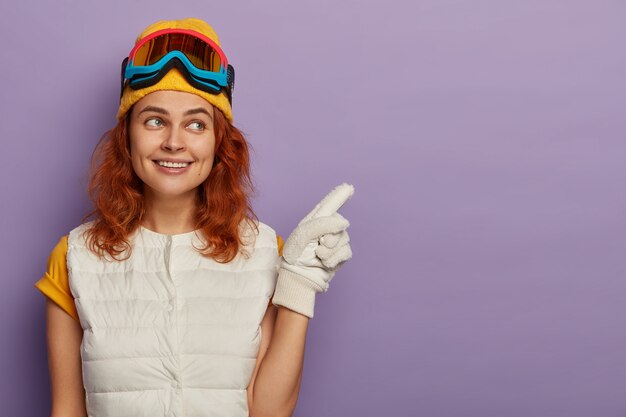 Jolie femme rousse porte un chapeau chaud, des lunettes de ski et un pull blanc