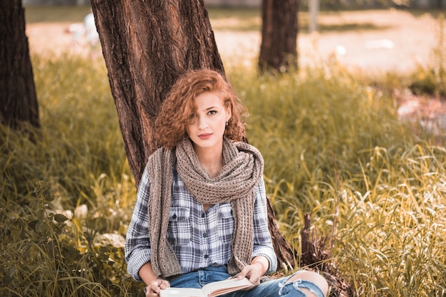 Photo gratuite jolie femme s'appuyant sur un arbre et tenant un livre dans un jardin public