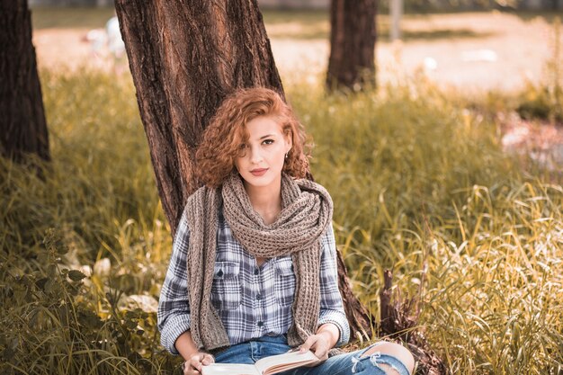 Jolie femme s&#39;appuyant sur un arbre et tenant un livre dans un jardin public