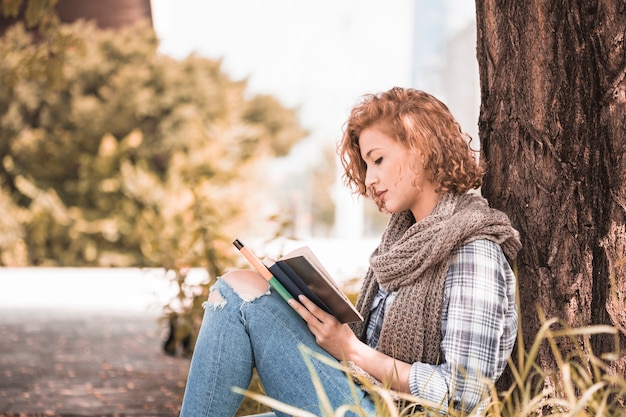 Jolie femme se penchant sur l&#39;arbre et livre de lecture dans un parc ensoleillé