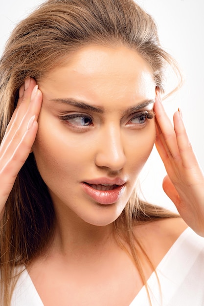 Jolie femme se tenant pour le visage et étirant la peau pour la faire paraître plus jeune Grandes lèvres yeux bleus en détournant les yeux Volume coiffure blonde maquillage naturel