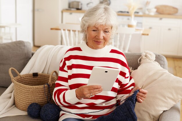 Jolie femme senior en sweat-shirt blanc rouge se détendre à l'intérieur, assis sur un canapé avec du fil et des aiguilles, tricoter, utiliser une tablette numérique pour les achats en ligne. Personnes âgées, retraite, technologie moderne
