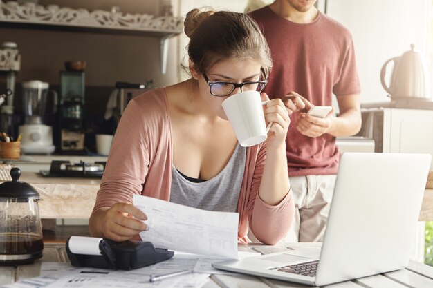 Jolie femme sérieuse à lunettes, boire du café et étudier le document dans ses mains, gérer le budget familial et faire de la paperasse à la table de la cuisine avec pile de factures, ordinateur portable et calculatrice