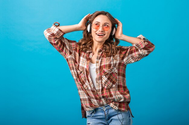 Jolie femme souriante appréciant écouter de la musique au casque en chemise à carreaux et jeans isolés sur fond bleu studio, portant des lunettes de soleil roses