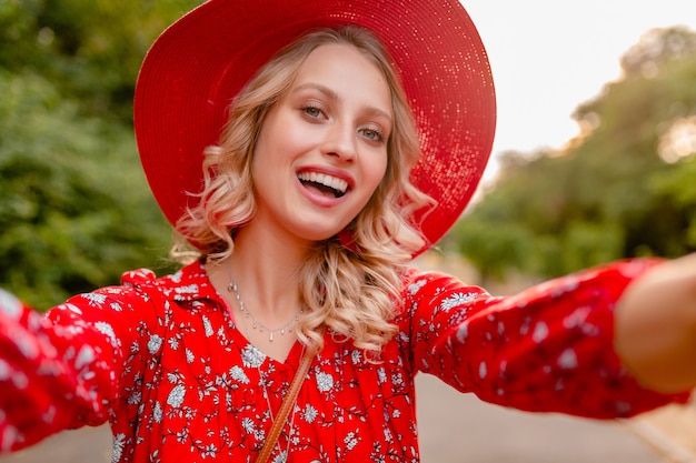 Jolie femme souriante blonde élégante en chapeau de paille rouge et blouse tenue de mode d'été prenant selfie photo