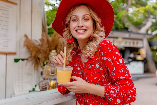 Jolie femme souriante blonde élégante en chapeau rouge paille et chemisier tenue de mode d'été buvant un smoothie cocktail de fruits naturels