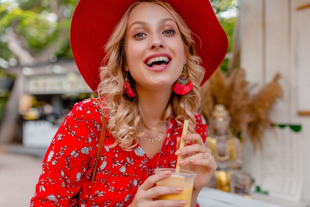 Jolie femme souriante blonde élégante en chapeau rouge paille et chemisier tenue de mode d'été buvant un smoothie cocktail de fruits naturels