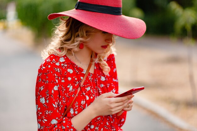 Jolie femme souriante blonde élégante en chapeau rouge de paille et tenue de mode d'été chemisier à l'aide de téléphone