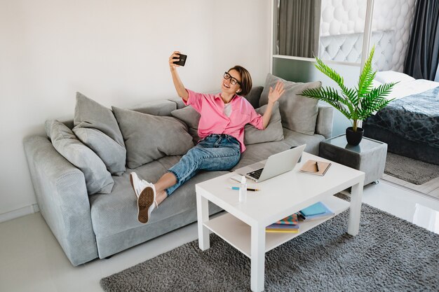 Jolie Femme Souriante En Chemise Rose Assise Détendue Sur Un Canapé à La Maison Dans Une Salle Intérieure Moderne à Table Travaillant En Ligne Sur Un Ordinateur Portable à La Maison