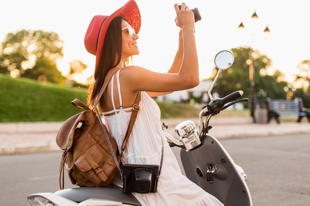 Jolie femme souriante à cheval sur la moto dans la rue en tenue de style estivale portant une robe blanche et un chapeau rouge voyageant avec sac à dos en vacances, prenant des photos sur un appareil photo vintage