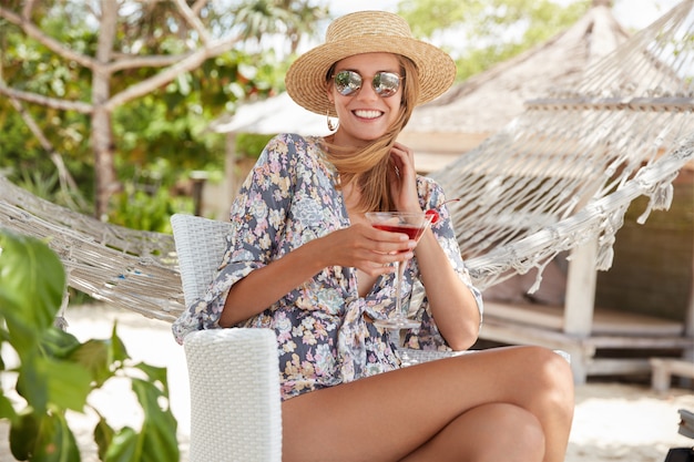 Jolie femme souriante dans des tons à la mode bénéficie d'un bon repos en plein air avec un cocktail frais, pose contre un hamac, heureuse de se retrouver entre amis et de faire une pause après le travail. Gens, loisirs et style de vie