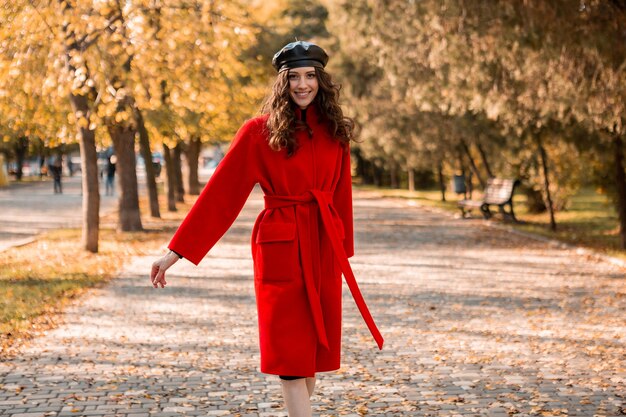 Jolie femme souriante élégante avec des cheveux bouclés marchant dans le parc habillé en manteau rouge chaud automne mode tendance, style de rue, portant un béret