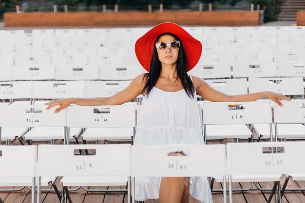 Jolie femme souriante heureuse habillée en robe blanche, chapeau rouge, lunettes de soleil assis dans le théâtre en plein air d'été sur une chaise seule, tendance de la mode de style de rue de printemps