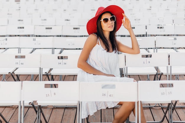 Jolie femme souriante heureuse habillée en robe blanche, chapeau rouge, lunettes de soleil assis dans le théâtre en plein air d'été sur une chaise seule, tendance de la mode de style de rue de printemps