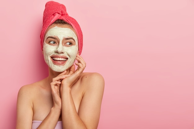 jolie femme souriante avec un masque d'argile, fait un pas de beauté, nettoie le visage, porte une serviette enveloppée sur la tête, se tient torse nu, obtient du plaisir, réduit les boutons, copie la zone de l'espace contre le mur rose