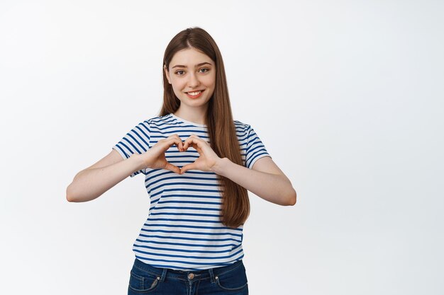 Jolie femme souriante montrant le signe du cœur près de la poitrine, debout sur fond blanc. Concept de relation et de romance