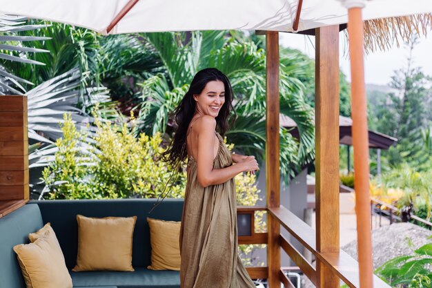 Jolie femme souriante positive européenne en robe de vol d'été à la lumière du jour naturelle à la terrasse de la villa bénéficiant de belles vacances, canapé extérieur avec oreillers sur tropical.