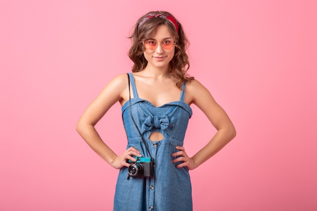 Jolie femme souriante prenant une photo sur un appareil photo vintage portant une robe en jean et des lunettes de soleil, isolé sur fond rose