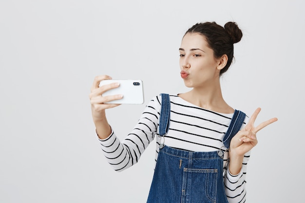 Jolie femme souriante prenant selfie sur smartphone, montrer le signe de la paix