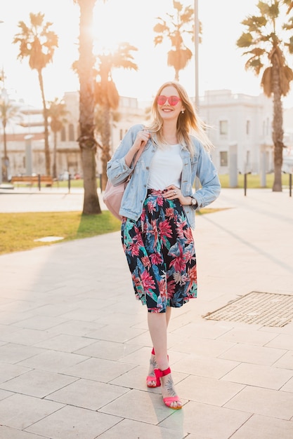 Jolie femme souriante qui marche dans la rue de la ville en jupe imprimée élégante et veste oversize en denim portant des lunettes de soleil roses, tendance de style d'été