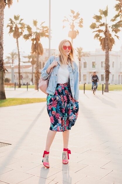 Jolie femme souriante qui marche dans la rue de la ville en jupe imprimée élégante et veste oversize en denim portant des lunettes de soleil roses, tendance de style d'été