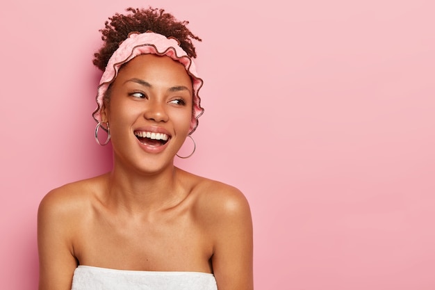 Jolie femme souriante regarde sur le côté droit se tient enveloppé dans une serviette avec des épaules nues porte un chapeau de douche aime prendre un bain et se détendre