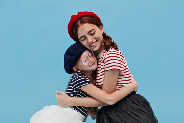 Jolie femme souriante en tenue rouge et noire étreignant avec sa petite soeur avec des taches de rousseur dans les lunettes sur fond bleu