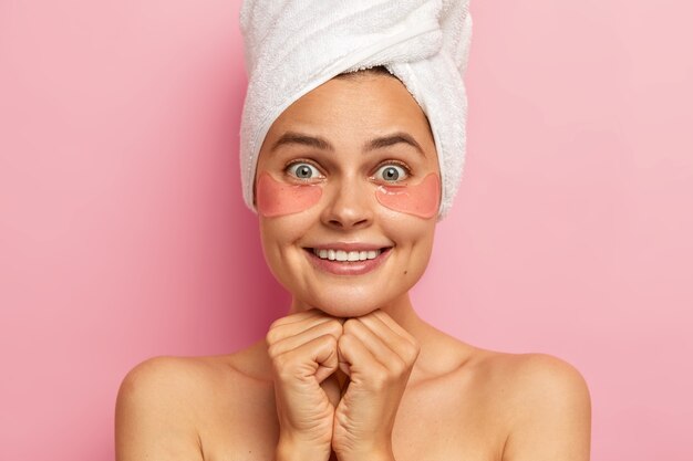 Jolie femme sourit agréablement, montre des dents blanches, applique des patchs sous les yeux pour réduire les rides