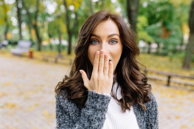 Jolie femme de style pose à la caméra dans le parc avec de vraies grandes émotions. Elle a l'air surprise et couvre son visage avec sa main et montre de vraies émotions.