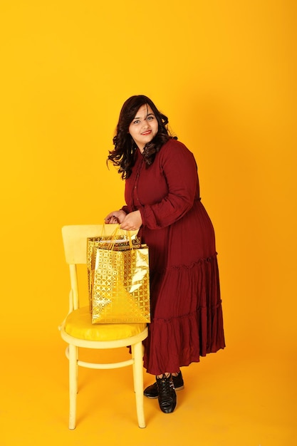 Photo gratuite jolie femme sud-asiatique en robe rouge foncé posée au studio sur fond jaune avec des sacs à provisions dorés