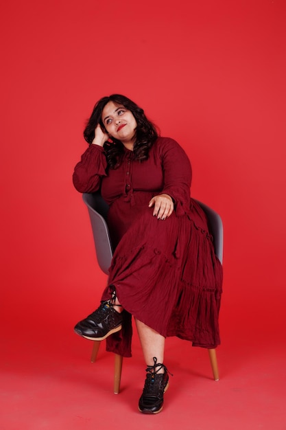 Jolie femme sud-asiatique en robe rouge foncé posée au studio sur fond rose assis sur une chaise