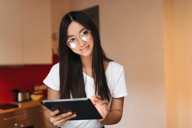 Jolie femme en T-shirt blanc et lunettes sourit, regardant devant et tenant la tablette