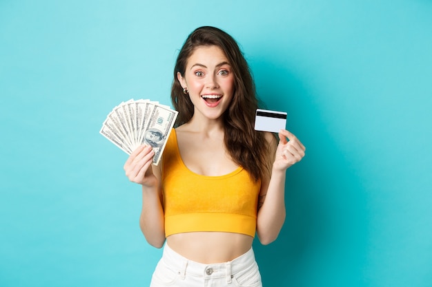 Jolie femme en tenue d'été, montrant des billets d'un dollar et une carte de crédit en plastique, souriante étonnée, debout sur fond bleu.