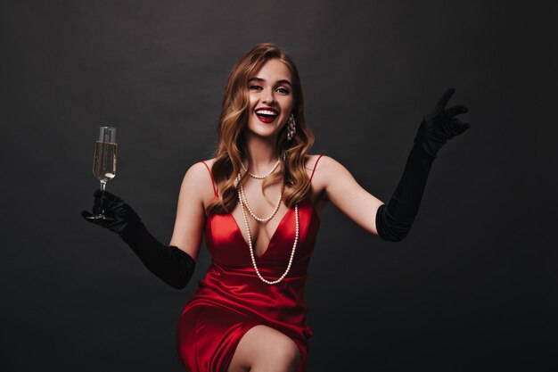 Jolie femme en tenue de soie et gants posant joyeusement sur fond noir et tenant du champagne