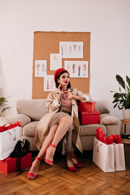 Jolie femme en trench-coat posant dans un appartement confortable Charmante jeune femme aux cheveux noirs en béret rouge est assise sur un canapé